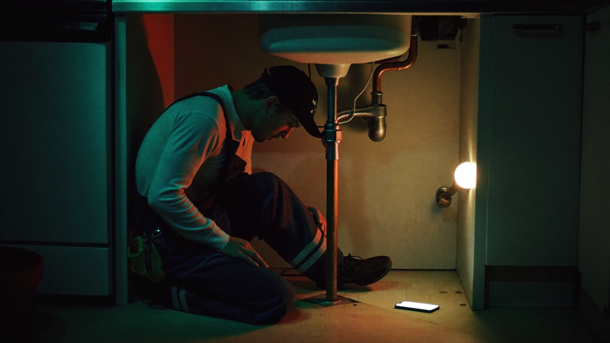 A plumber kneels under a kitchen sink at night, fixing pipes, while his smartphone glows on the floor beside him